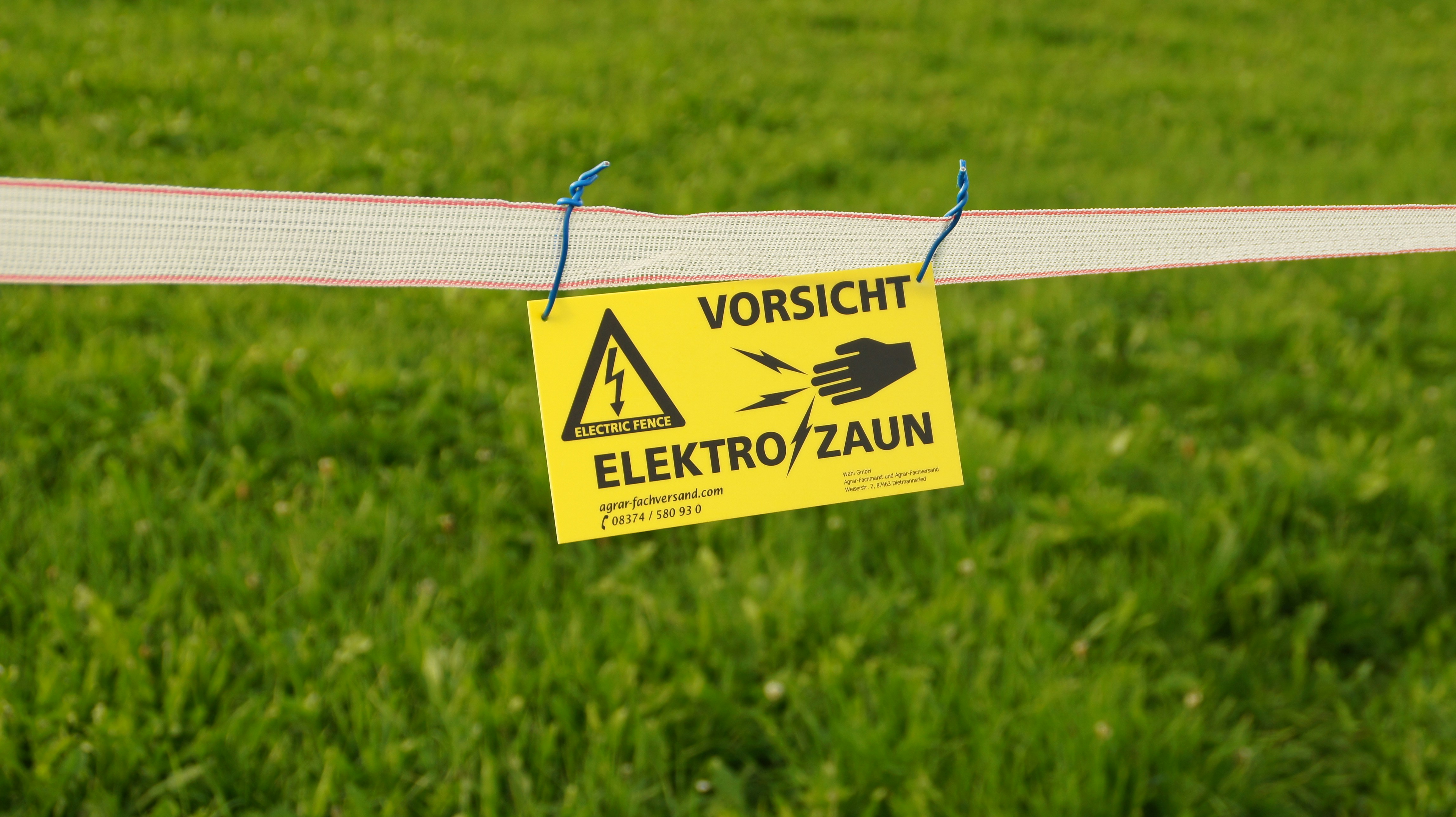 A vibrant yellow warning sign hangs on an electric fence in the middle of a lush green field. The sign prominently displays a cautionary message in German, warning of electric shocks with clear symbols for safety. This setup is typical in rural or agricultural areas to protect fields or livestock. The contrasting colors make the sign highly visible, ensuring effective communication of potential danger. The serene countryside contrasts with the functional and necessary safety precautions.